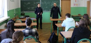 W klasie szkolne widać umundurowanego policjanta, umundurowaną policjantkę, przed nimi ławki uczniowskie, w których siedzą uczniowie.