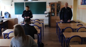 W klasie szkolne widać umundurowanego policjanta, umundurowaną policjantkę, przed nimi ławki uczniowskie, w których siedzą uczniowie.