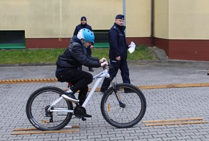 na zewnątrz  budynku szkoły chłopiec jedzie na rowerze w kasku za nim dwóch umundurowanych policjantów