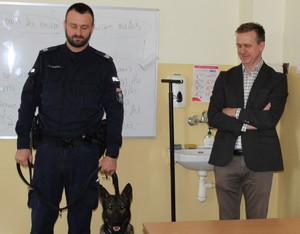 w pomieszczeniu budynku szkołu, w klasie stoi jeden umundurowany policjant,  obok niego na na pies oraz mężczyzna