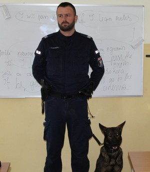 w pomieszczeniu budynku szkoły, w klasie szkolnej stoi umundurowany policjant obok niego siedzi  duży pies o