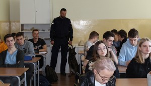 w pomieszczeniu budynku szkoły, klasa uczniowska stoi umundurowany policjant obok niego duży pies, przed nimi siedzą za stolikami uczniowie