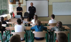 W pomieszczeniu, w klasie uczniowskiej stoi jeden umundurowany policjant, jedna umundurowana policjantka, przed nimi w ławkace siedzi kilkoro uczni.