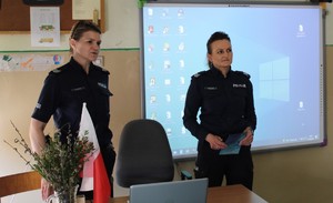 W pomieszczeniu, w klasie uczniowskiej stoją dwie umundurowane policjantki.
