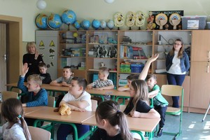 W pomieszczeniu, w klasie uczniowskiej stoją dwie nauczycielki przed nimi w ławkach siedzi kilkoro uczni.