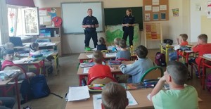 W klasie szkolnej umundurowany policjant, umundurowana policjantka, przed nimi w ławkach szkolnych siedzi kilkanaście dzieci.