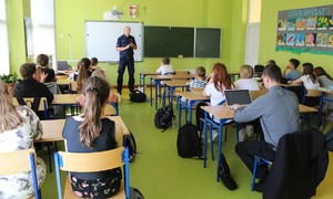 W klasie szkolnej jeden umundurowany policjant, w szkolnych ławkach siedzi kilkanaście dzieci.