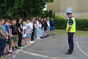 Boisko szkolne. Grupa dzieci stojąca w rzędzie, przed  nimi policjant.