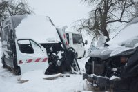 Na zdjęciu widoczne są trzy pojazdy typu bus, dwa koloru czarnego są zasypane śniegiem i posiadają uszkodzone i zdekompletowane przody. Na jednym z czarnych busów oparte sa drzwi koloru białego z biało czerwoną naklejką. W oddali widać tylko część białego busa, który stoi za czarnymi busami.