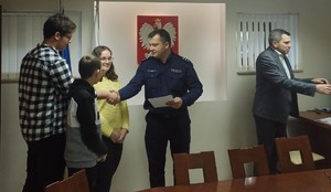 W pomieszczeniu budynku urzędu stoi jeden umundurowany policjanty, mężczyzna, dwóch  chłopców, jedna dziewczynka, która trzyma papierowy dyplom w postaci kartki papieru formatu A4, umundurowany policjant ściska dłoń jednego z chłopców, gratuluje.