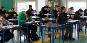 W pomieszczeniu szkoły w klasie uczniowskiej kilkanaście  uczni siedzi pojedynczo w ławkach, rozwiązują test.