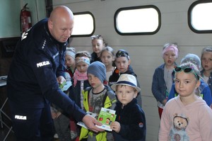 W pomieszczeniu umundurowany policjant wręcza dziecku ulotkę, obok kilkoro dzieci.