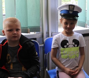 jedna dziewczynka w czapce policyjnej z ruchu drogowego, jeden chłopiec, dzieci siedzą w sali