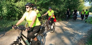 Dwie kobiety na drodze leśnej jedzie rowerami za nimi jadą dwóch umundurowanych policjantów.