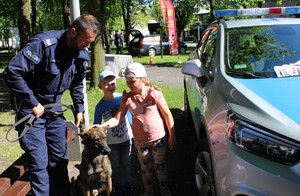 Na zewnątrz umundurowany policjant trzyma na smyczy psa, obok dwoje dzieci, obok część radiowozu oznakowanego.