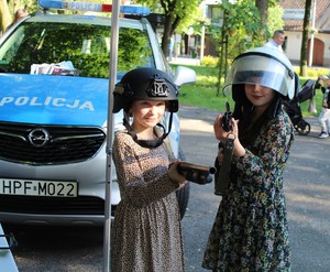 Na zewnątrz dwie dziewczynki, które na głowach mają kaski policyjne, w ręku trzymają broń, za nimi widać część oznakowanego radiowozu.