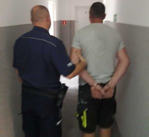 Na korytarzu umundurowany policjant, który trzyma mężczyznę zapiętego w kajdanki na ręce założone w tyle. Obaj mężczyźni odwróceni plecami.