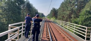 Na trakcji kolejowej funkcjonariusz umundurowany SOK lornetką obserwuje otoczenie, obok umundurowany policjant. Oboje odwróceni tyłem.