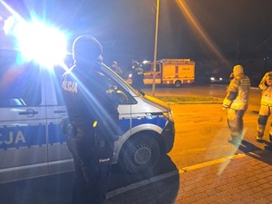 noc, radiowóz oznakowany policyjny, obok policjant w mundurze, w oddali dwóch umundurowanych strażaków