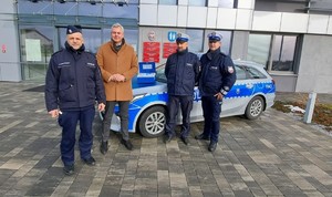 Przed budynkiem zaparkowany oznakowany radiowóz, przed nim stoją 3 umundurowanych policjantów i mężczyzna.