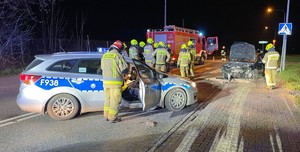 Zdjęcie opisujące zdarzenie drogowe. Jest ciemno, radiowóz, wóz strażacki, rozbity samochód osobowy, kilku strażaków.