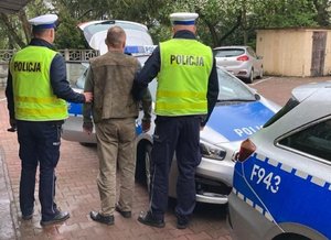 Dwóch umundurowanych policjantów trzyma mężczyznę z kajdankami zapiętymi na ręce trzymane w tyle.