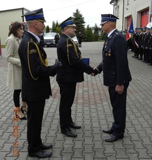 Trzech strażaków. Dwóch z nich podaje sobie prawe dłonie.