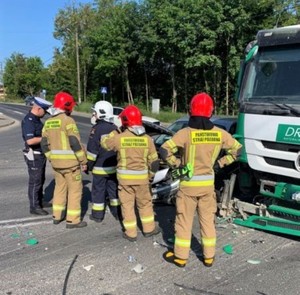 Na zdjęciu zdarzenie drogowe osobówki i tira. Obok uszkodzonych pojazdów strażacy i policjant.