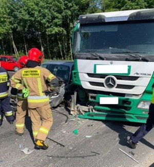 Na zdjęciu zdarzenie drogowe osobówki i tira. Obok uszkodzonych pojazdów strażacy i policjant.