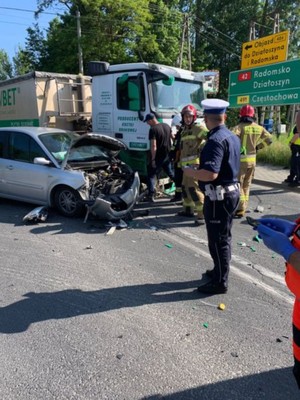 Na zdjęciu zdarzenie drogowe osobówki i tira. Obok uszkodzonych pojazdów strażacy i policjant.