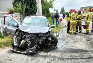 Na drodze rozbity samochód osobowy, obok grupa strażaków i policjant.