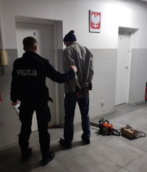 Policjant i zatrzymany mężczyzna stoją odwróceni tyłem. Mężczyzna kajdanki na rękach zapiętych w tyle. Obok na podłodze leżą piła i prostownik samochodowy.