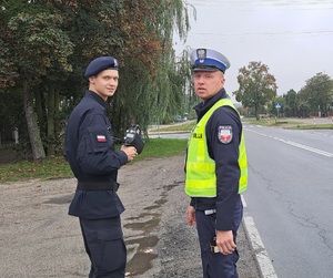 Funkcjonariusz i uczeń klasy mundurowej stoją na poboczu drogi.