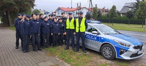 Dwóch policjantów i grupa uczniów w mundurach szkolnych  stoją przy radiowozie.