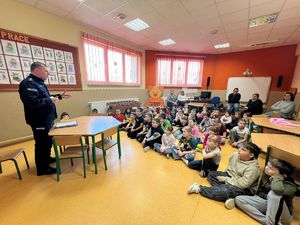 Umundurowany policjant stoi przed grupą siedzących w ławkach dzieci.
