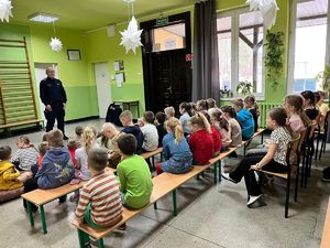 Umundurowany policjant stoi przed grupą siedzących w ławkach dzieci.