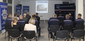 Umundurowany policjant występuje, przed nimi grupa siedzących osób i grupa umundurowanych policjantów.