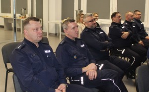 6 umundurowanych policjantów siedzi na krzesłach, za nimi siedzą dwie kobiety.