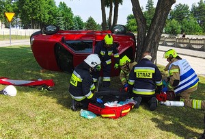 Trzech strażaków reanimuje osobę poszkodowaną w wypadku, obok na boku, przy drzewie samochód osobowy.