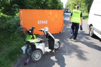 Rozbity wózek inwalidzki, pomarańczowy parawan z napisem Straż oraz policjant wykonujacy czynnosci na miejscu zdarzenia.